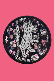A round black velcro patch with an embroidered design of a wolf and floral pattern, and the text 'Woolf With Me'.