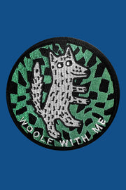 A round embroidered patch depicting a wolf's face in a checkered pattern on a green background, with the text 'Woolf With Me' below it.