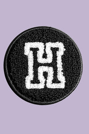 Circular patch with a white letter 'H' on a black background, against a light purple background