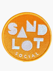 Woolf With Me® round orange embroidered Velcro patch with Sandlot Social text design on a wooden surface