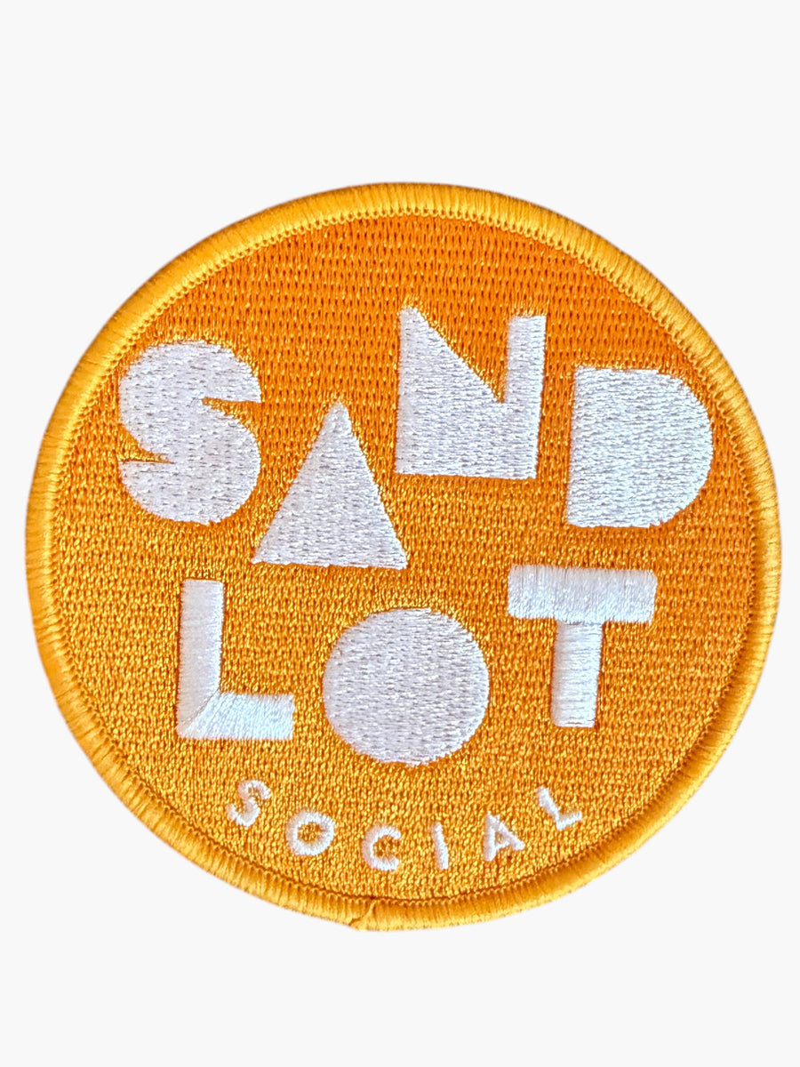 Woolf With Me® round orange embroidered Velcro patch with Sandlot Social text design on a wooden surface